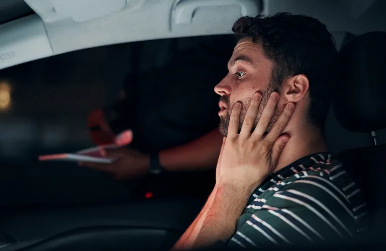 Young man in striped shirt sitting in car, holding his face with both hands, speaking to a person outside at night.
