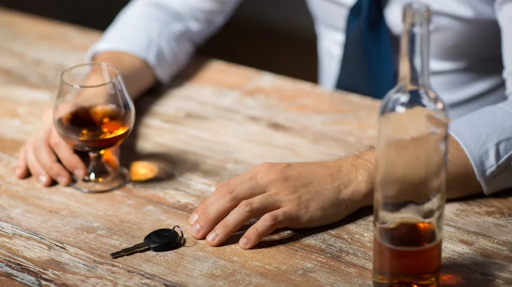 Man in white shirt and blue tie holding a glass of whiskey, car key and bottle on wooden table.