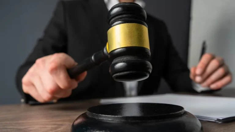 Person in a dark suit holding a black and gold judge's gavel over a wooden desk.