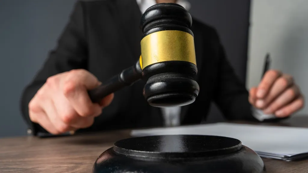 Person in a dark suit holding a black and gold judge's gavel over a wooden desk.