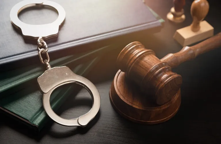 Metal handcuffs resting on closed books beside a wooden judge's gavel on a dark surface.