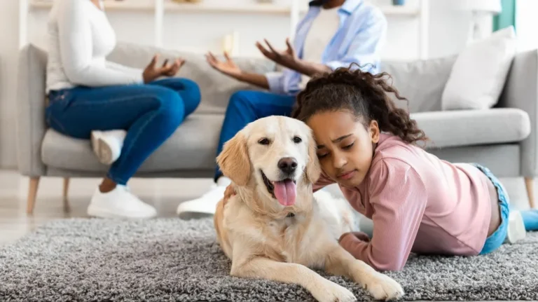 Young girl lying on gray carpet next to golden retriever dog, two adults conversing on gray sofa in background.