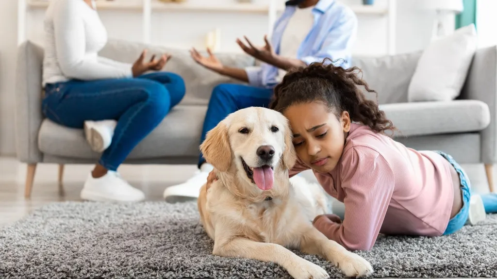 Young girl lying on gray carpet next to golden retriever dog, two adults conversing on gray sofa in background.
