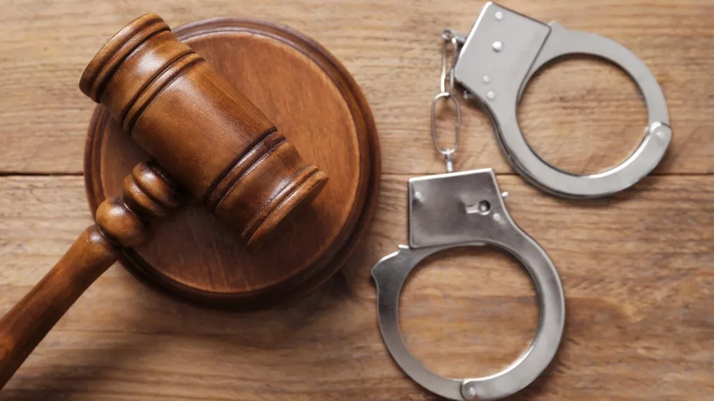 Wooden judge's gavel and round sound block beside metallic handcuffs on wooden surface.