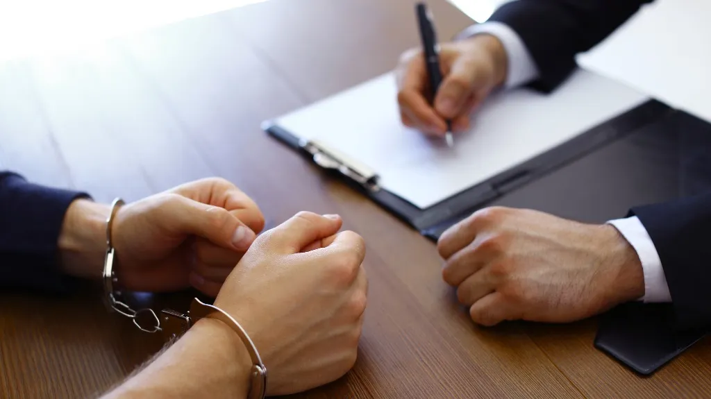 Person in handcuffs seated across a table from a professional taking notes on a clipboard.