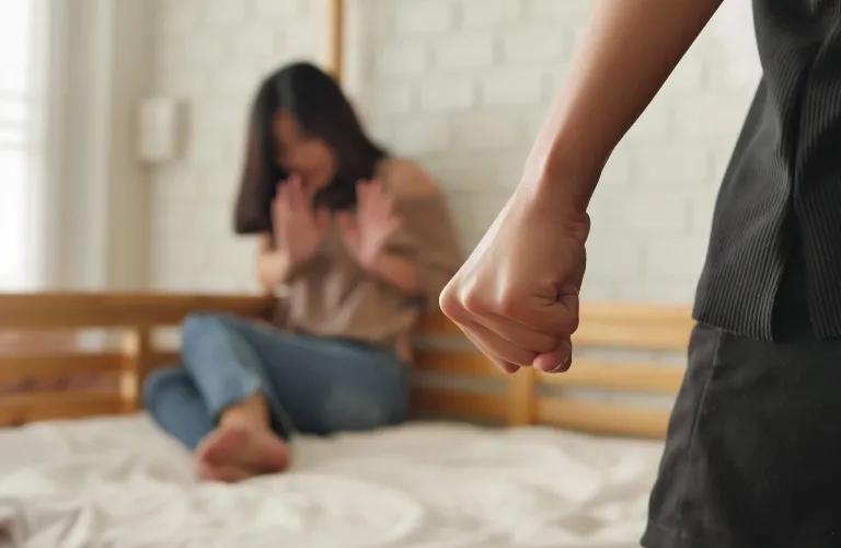 Person with clenched fist in foreground, woman sitting on bed with raised hands in defensive posture.
