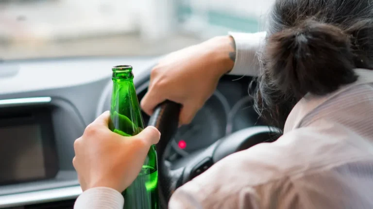 Person holding a green glass bottle while gripping a car steering wheel.