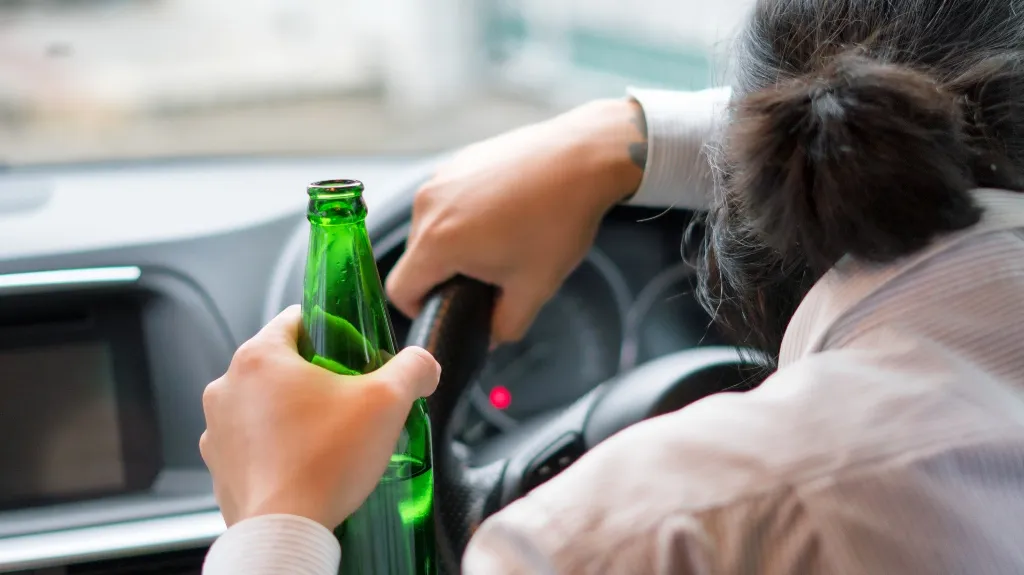 Person holding a green glass bottle while gripping a car steering wheel.
