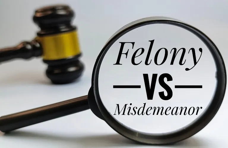 Magnifying glass focusing on the words "Felony vs Misdemeanor" with a wooden gavel in the background.