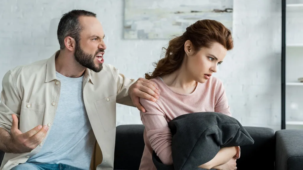 Middle-aged man angrily shouting at woman holding a pillow, both seated on a sofa.