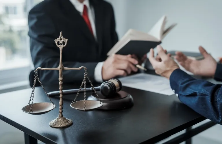 Bronze scales of justice and wooden gavel on table with two professionals discussing legal documents.