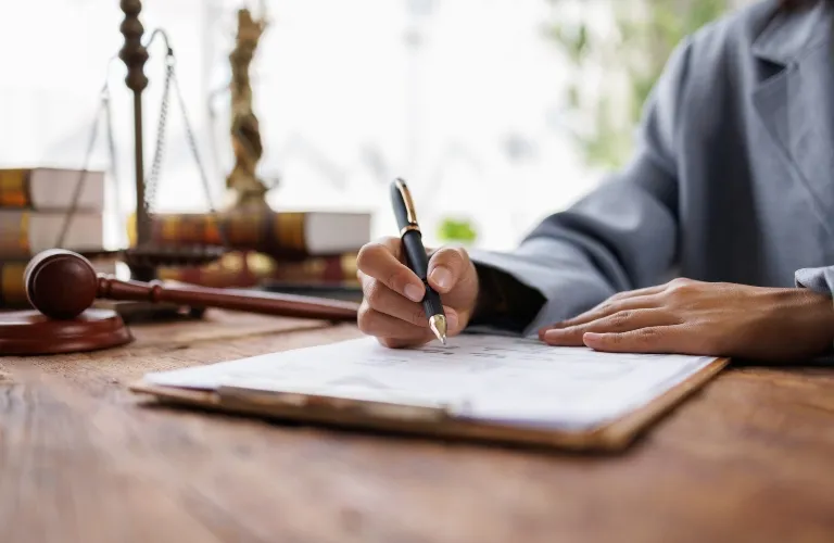 Person in gray suit writing on clipboard at wooden desk with gavel and law books.