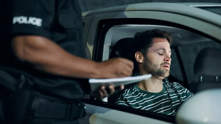 Police officer writing a ticket for a man in a green striped shirt sitting inside a white car.