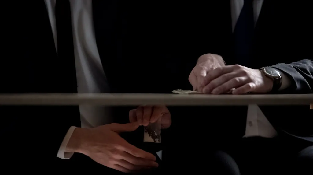 Two individuals in dark suits exchanging a small plastic bag and cash over a table in low light.
