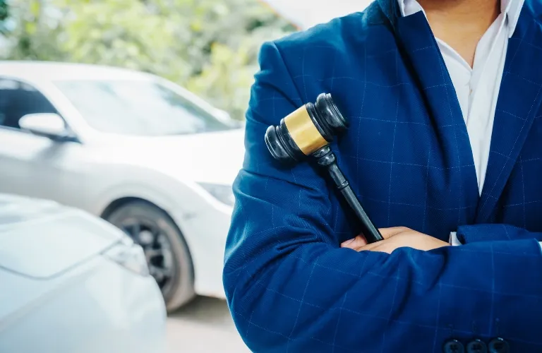 Person in blue suit holding a black and gold gavel with crossed arms, cars in background.