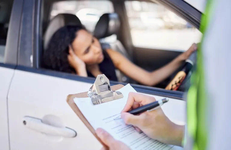 Woman sitting in white car with hand on head while officer writes on clipboard outside vehicle.