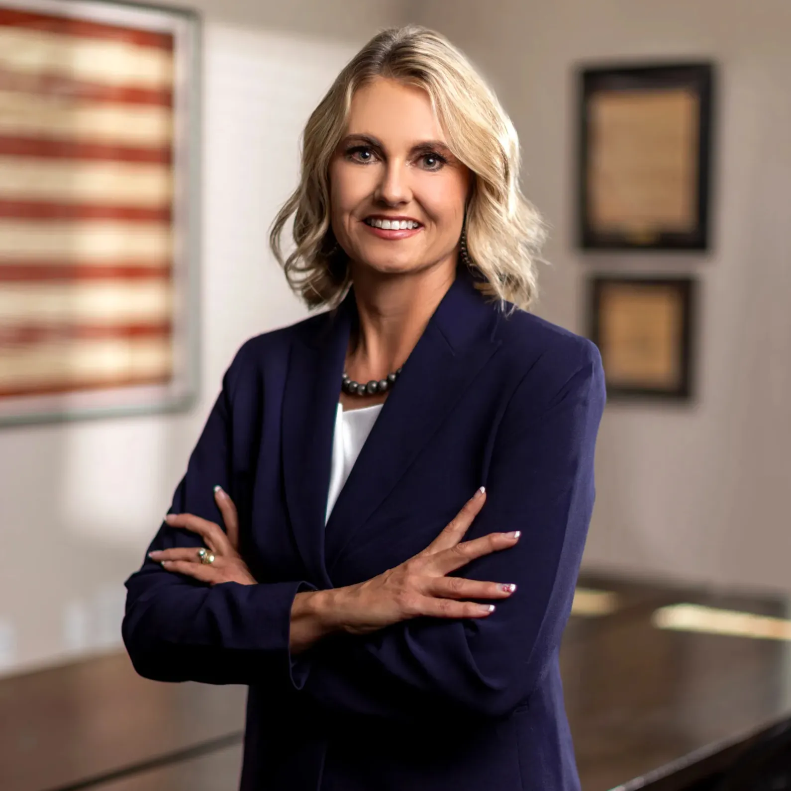 Professional woman with blonde hair wearing navy blazer and pearl necklace, standing with arms crossed.