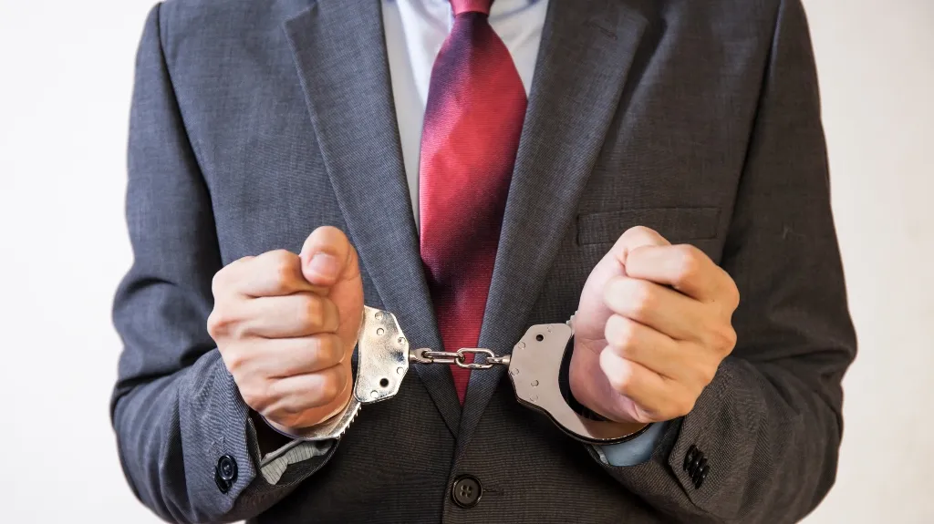 Man in gray suit and red tie with hands cuffed in silver handcuffs.