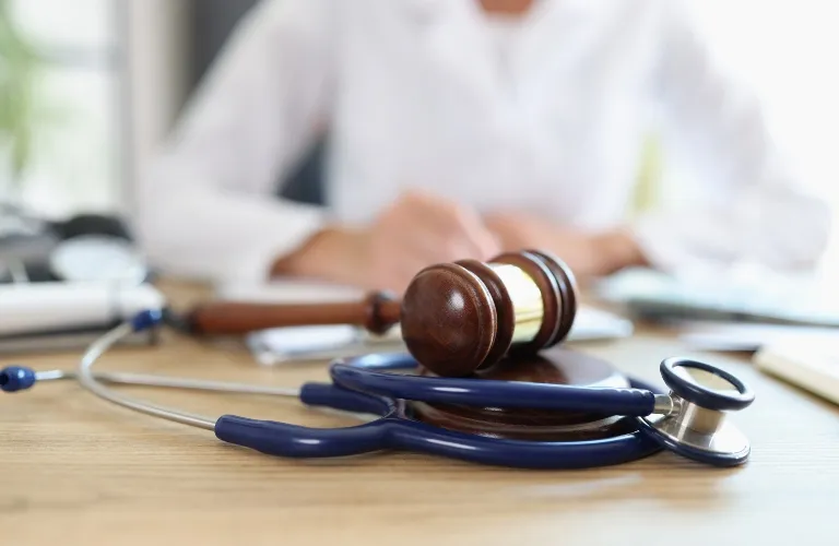 Wooden judge’s gavel resting on a desk next to a blue stethoscope, with a blurred person in white in the background.