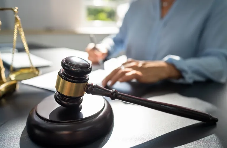 Wooden judge's gavel with brass band on desk, person writing in background.