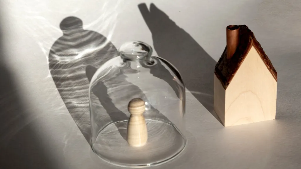 Wooden pawn figure under a glass dome next to a small wooden house model on a white surface.