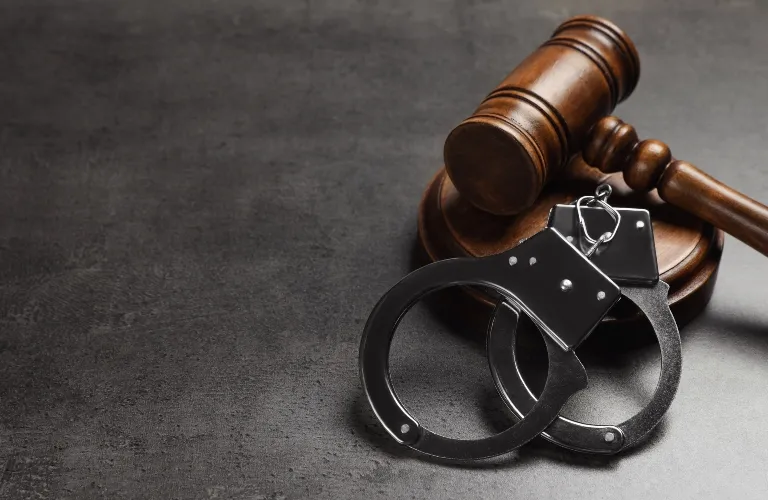 Wooden judge's gavel resting on block beside black metal handcuffs on gray surface.