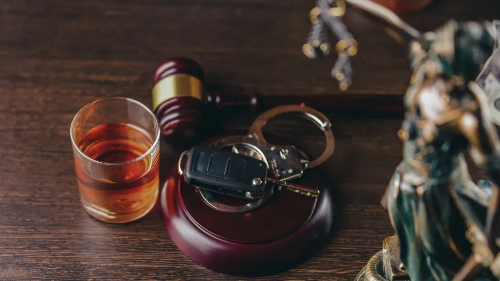 Glass of amber liquid, wooden judge's gavel, car keys, and handcuffs on dark wood surface.