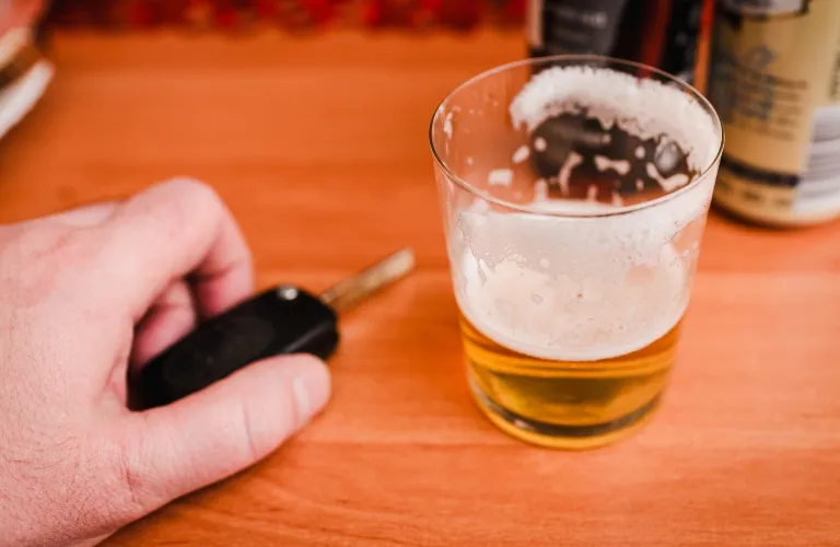 Hand holding a car key next to a partially filled glass of beer on a wooden surface.
