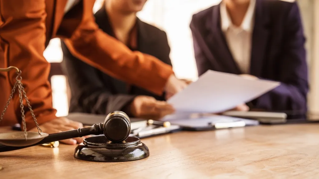 Wooden judge's gavel and scales on table with three business professionals reviewing documents.