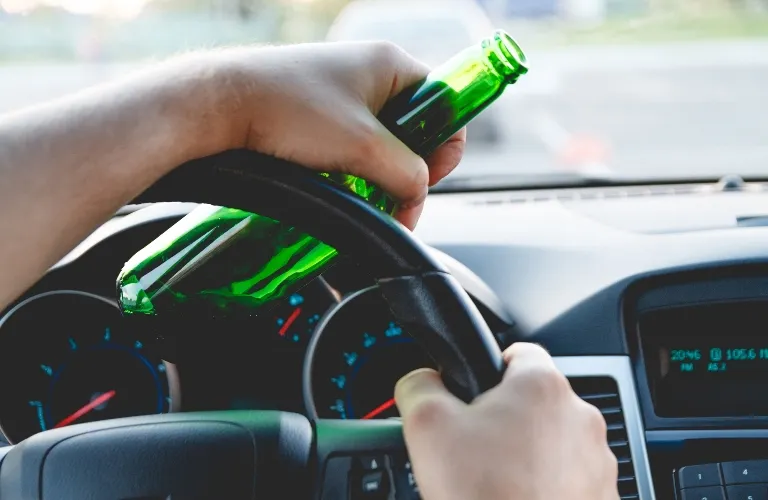 Person holding a green glass bottle while gripping a car steering wheel inside a vehicle.
