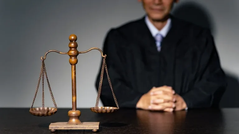 Bronze balance scale in focus with a judge in a black robe sitting behind a dark wooden table.