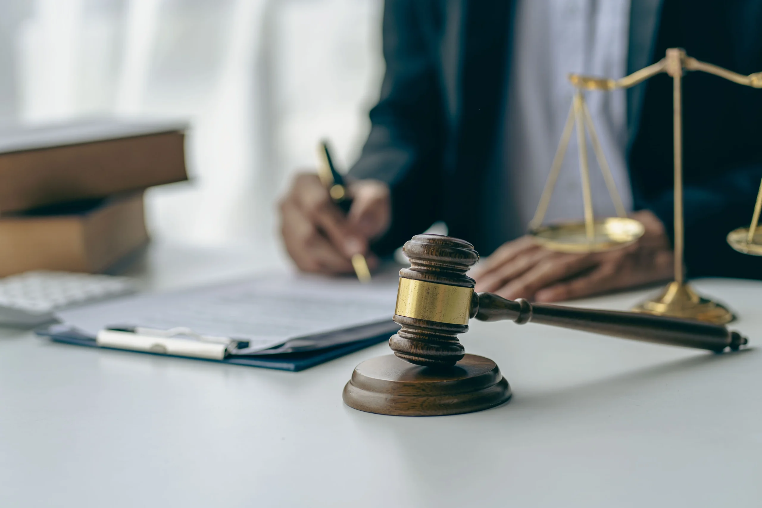 Wooden judge's gavel, brass scales of justice, and person signing documents in background.