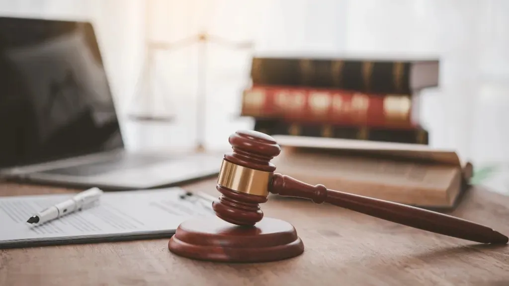 Wooden judge's gavel resting on a sound block with legal books, documents, and a laptop.